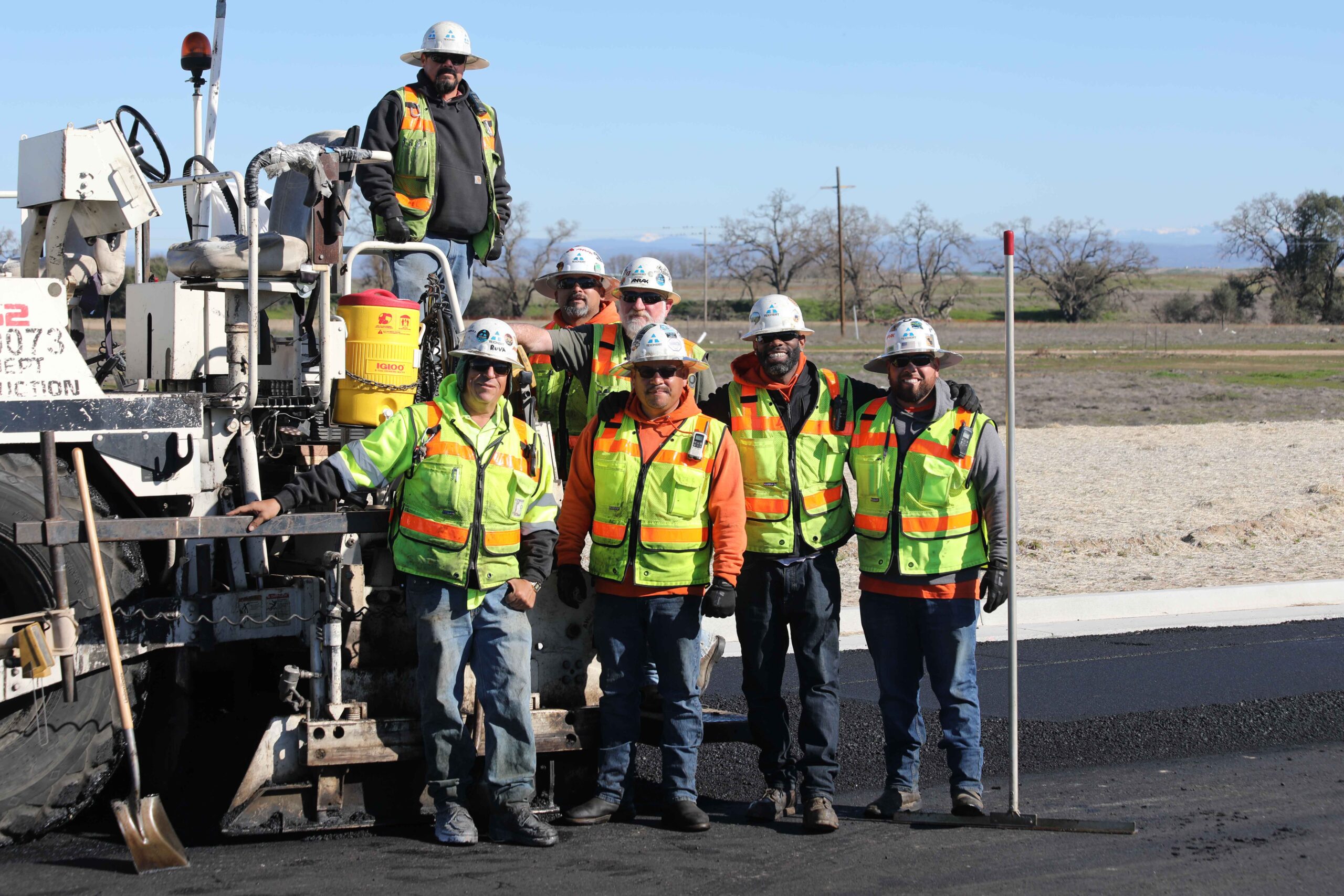 Teichert Construction workers all wearing face coverings and orange vests while standing in front of a Teichert green construction vehicle